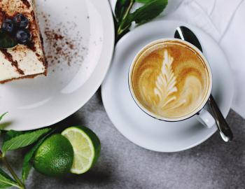 A cup of coffee on a table next to a slice of cake and a lime